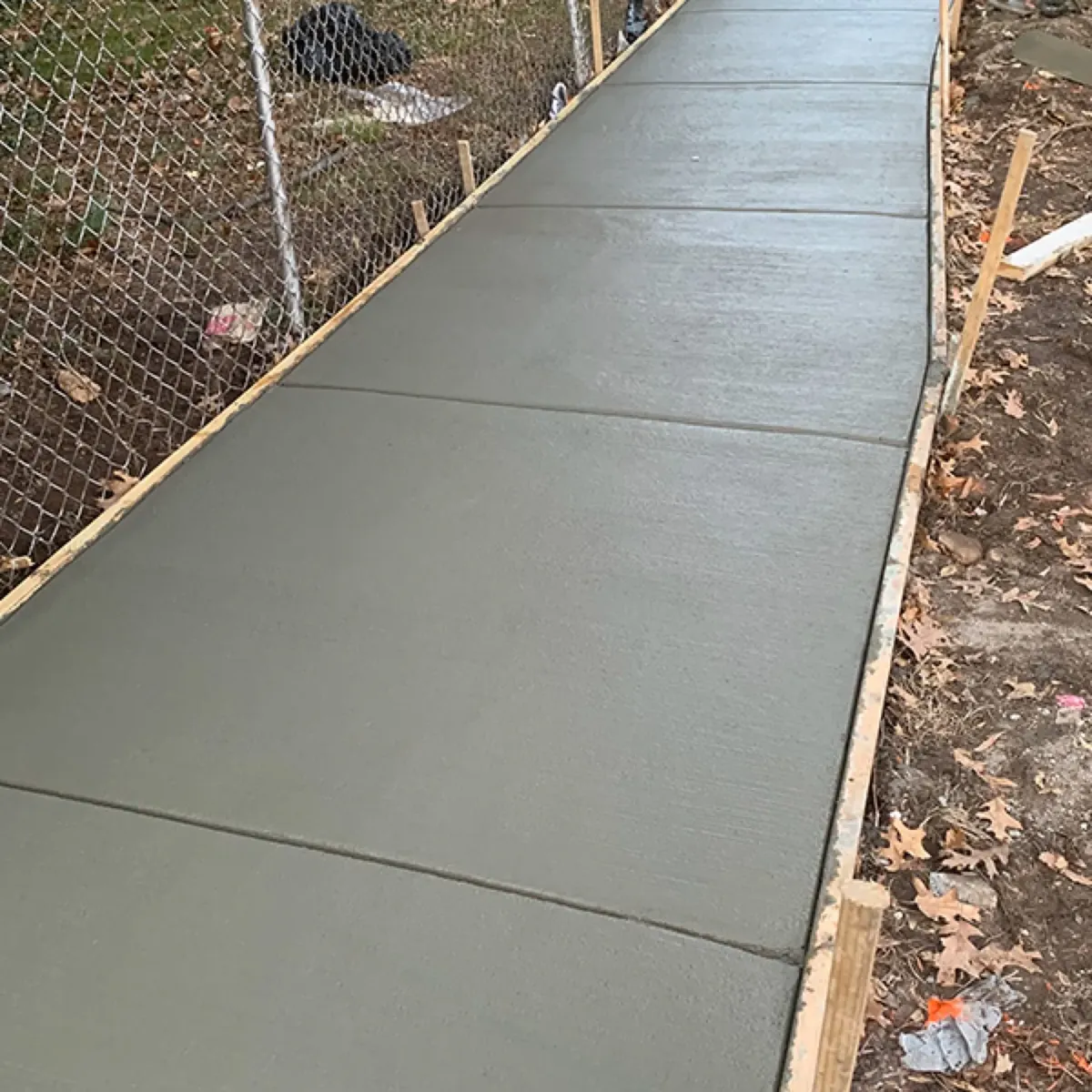 Freshly poured narrow concrete walkway in wood forms with tooled joints dividing the panels along a fence
